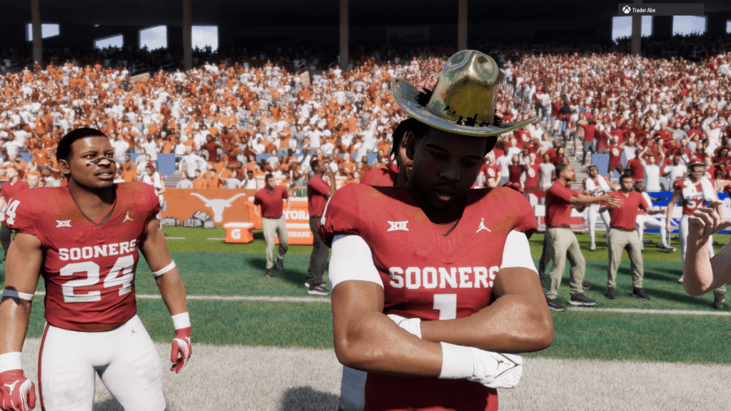Oklahoma Sooners running back Isaac Brown stands on the field after the Red River Rivalry win over Texas, wearing a gold cowboy hat with his arms crossed, while teammate Xavier Robinson smiles beside him amid a split crowd of crimson and burnt orange fans.